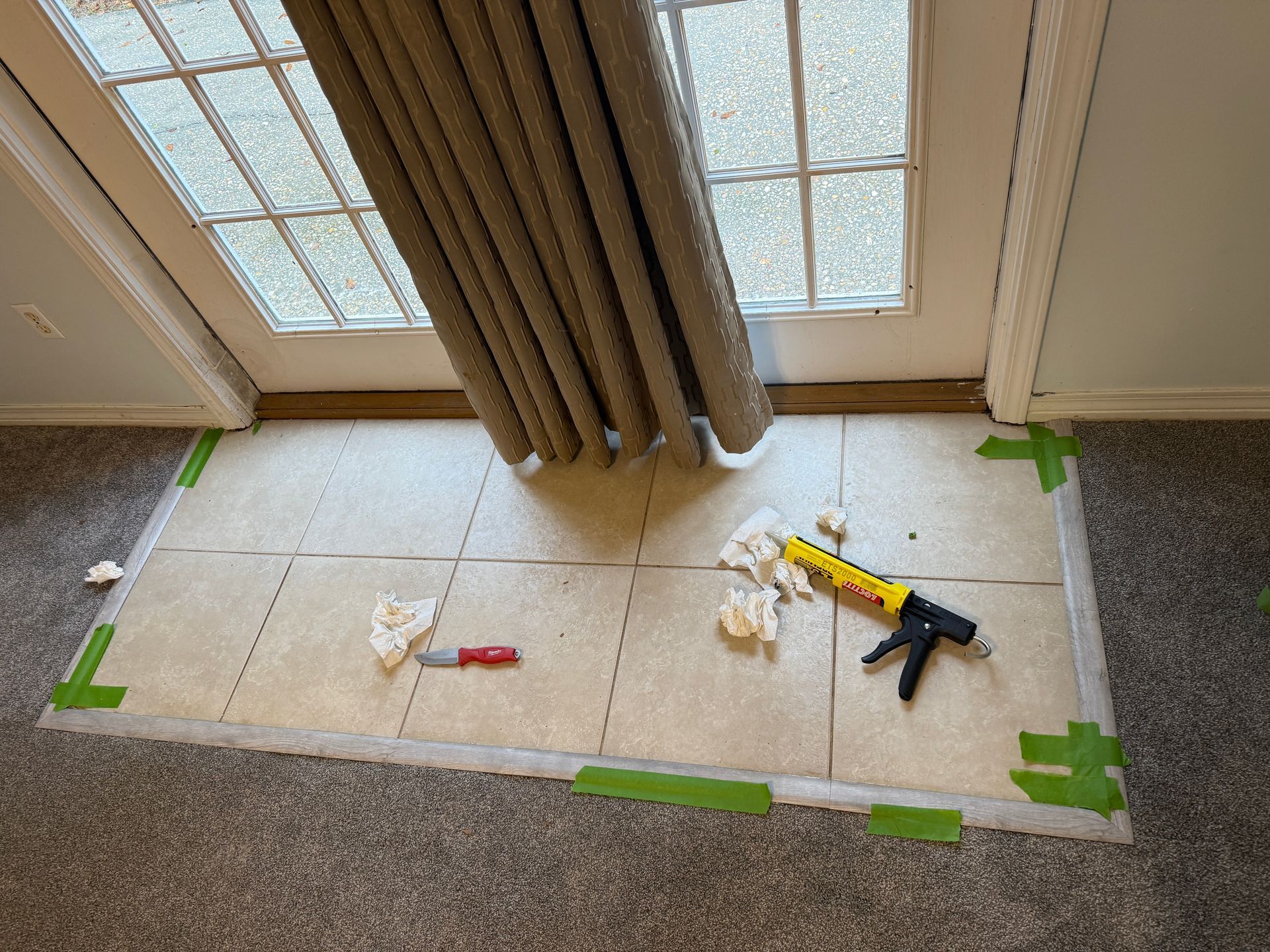 Tiled floor area by a door, with caulk gun, knife, tape, and curtain.