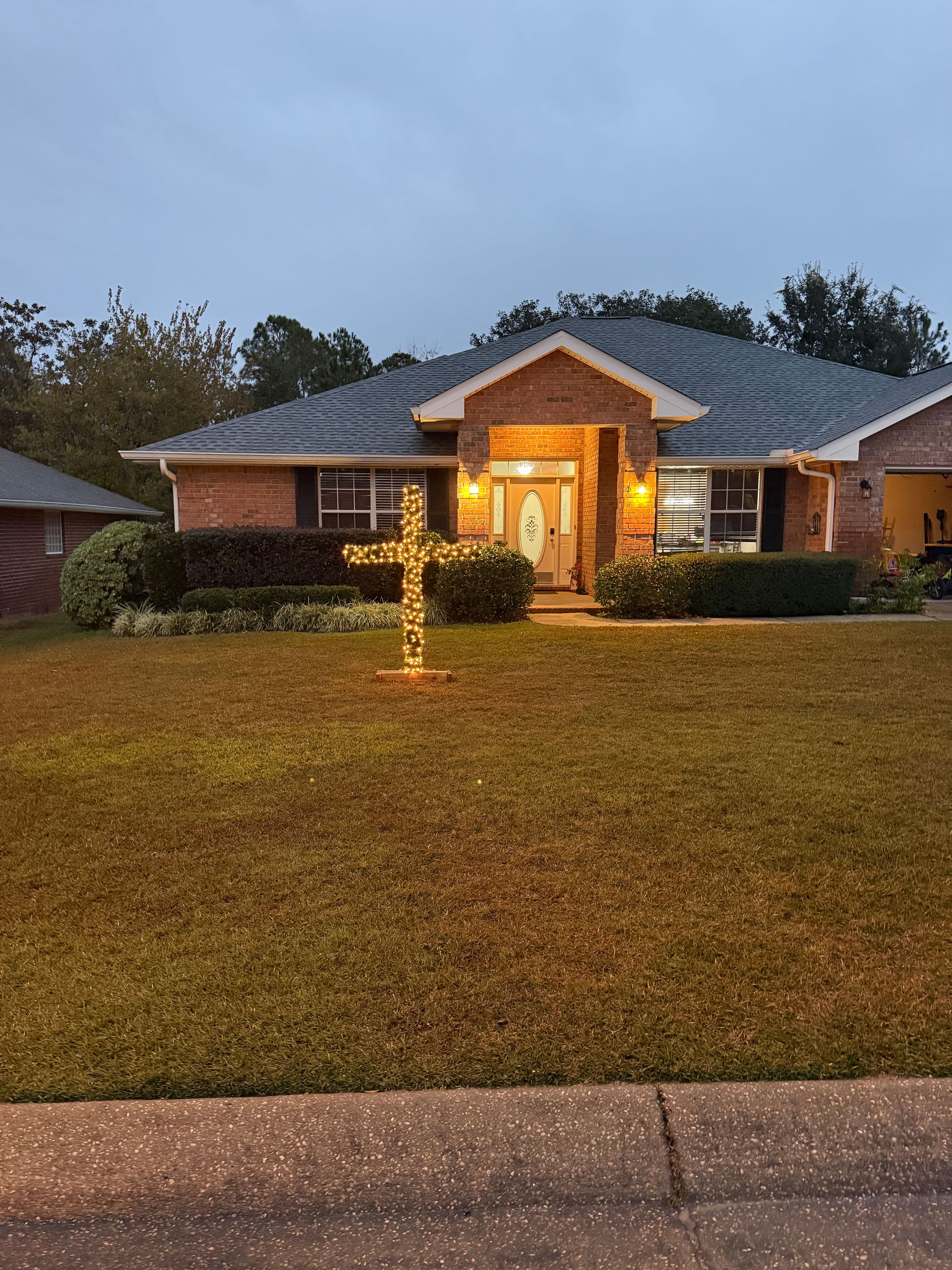 Brick house with glowing cross in front yard at dusk.