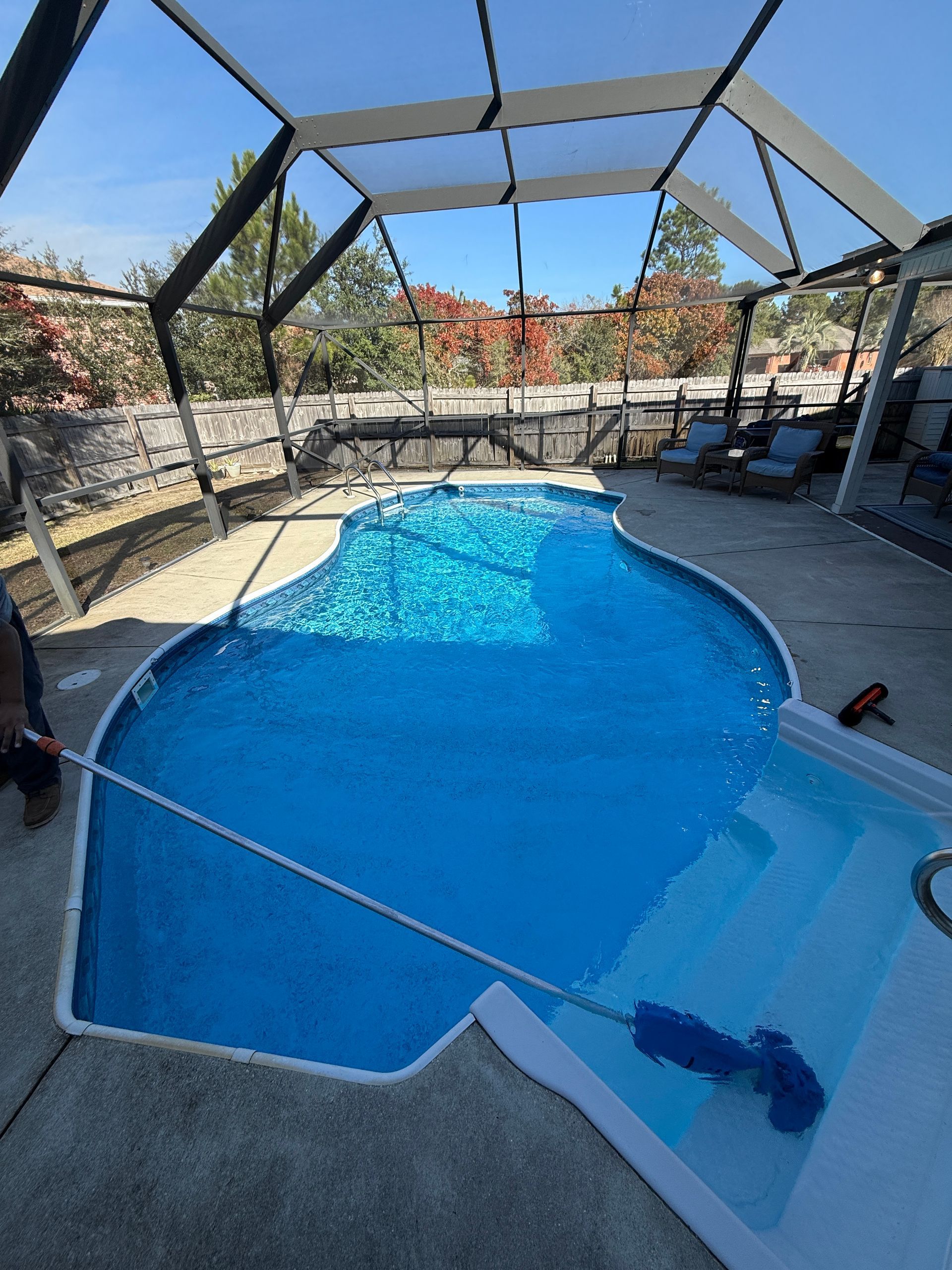 A pool with blue water under a screened enclosure. A person is cleaning the pool with a brush.
