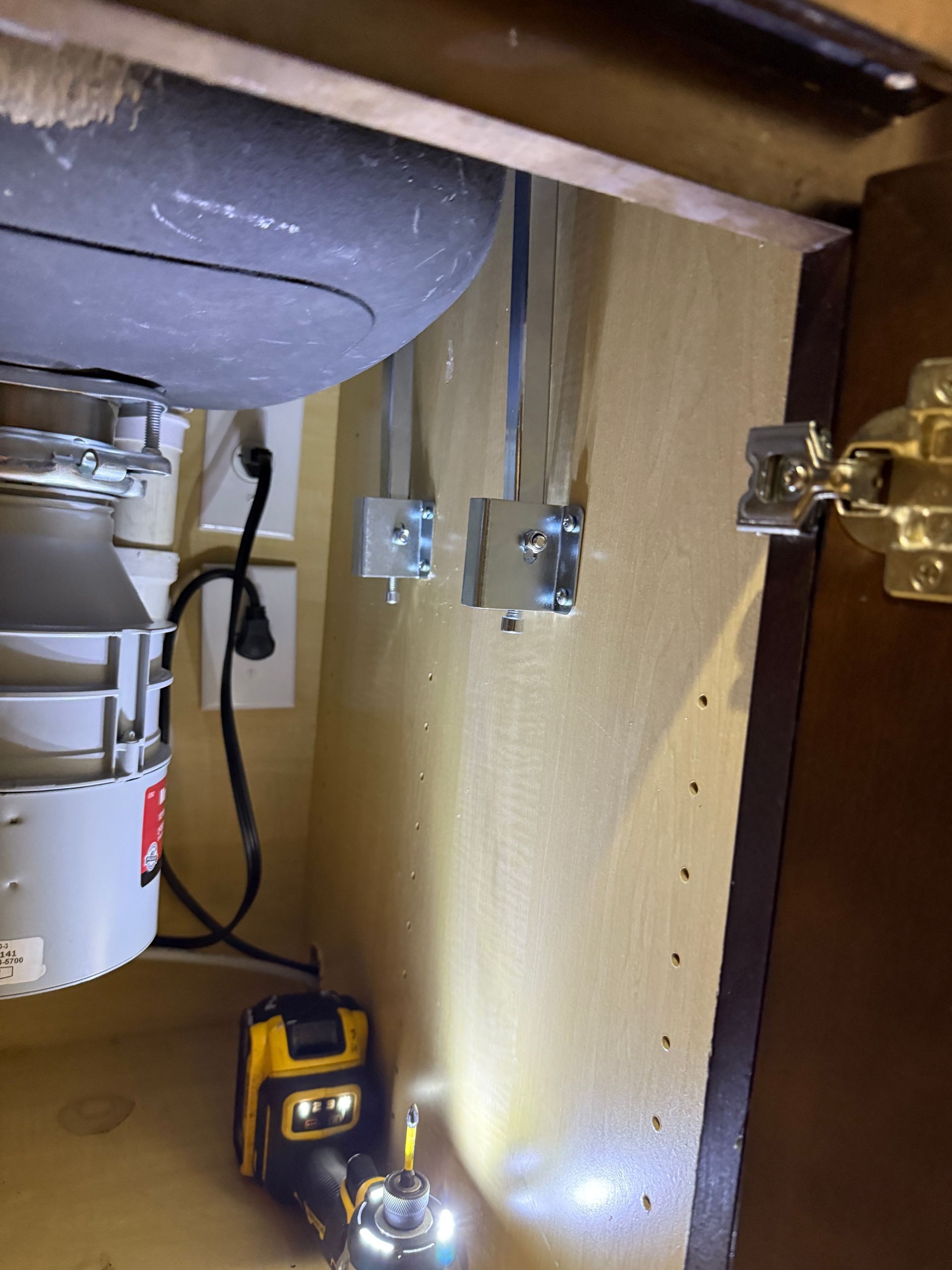 Under-sink cabinet with garbage disposal, electrical outlets, and metal cable mounts on the wall. A drill rests on the shelf.