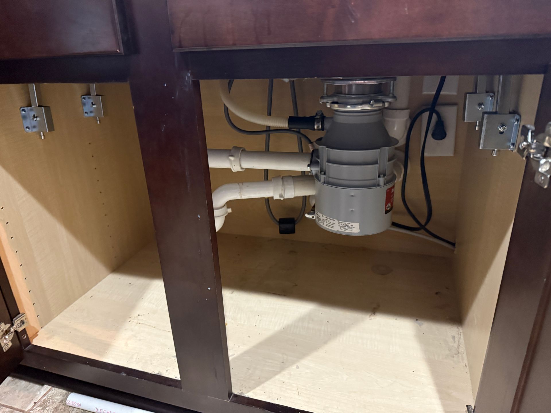 Under-sink cabinet with a garbage disposal and plumbing visible. Brown cabinet with white pipes and disposal.