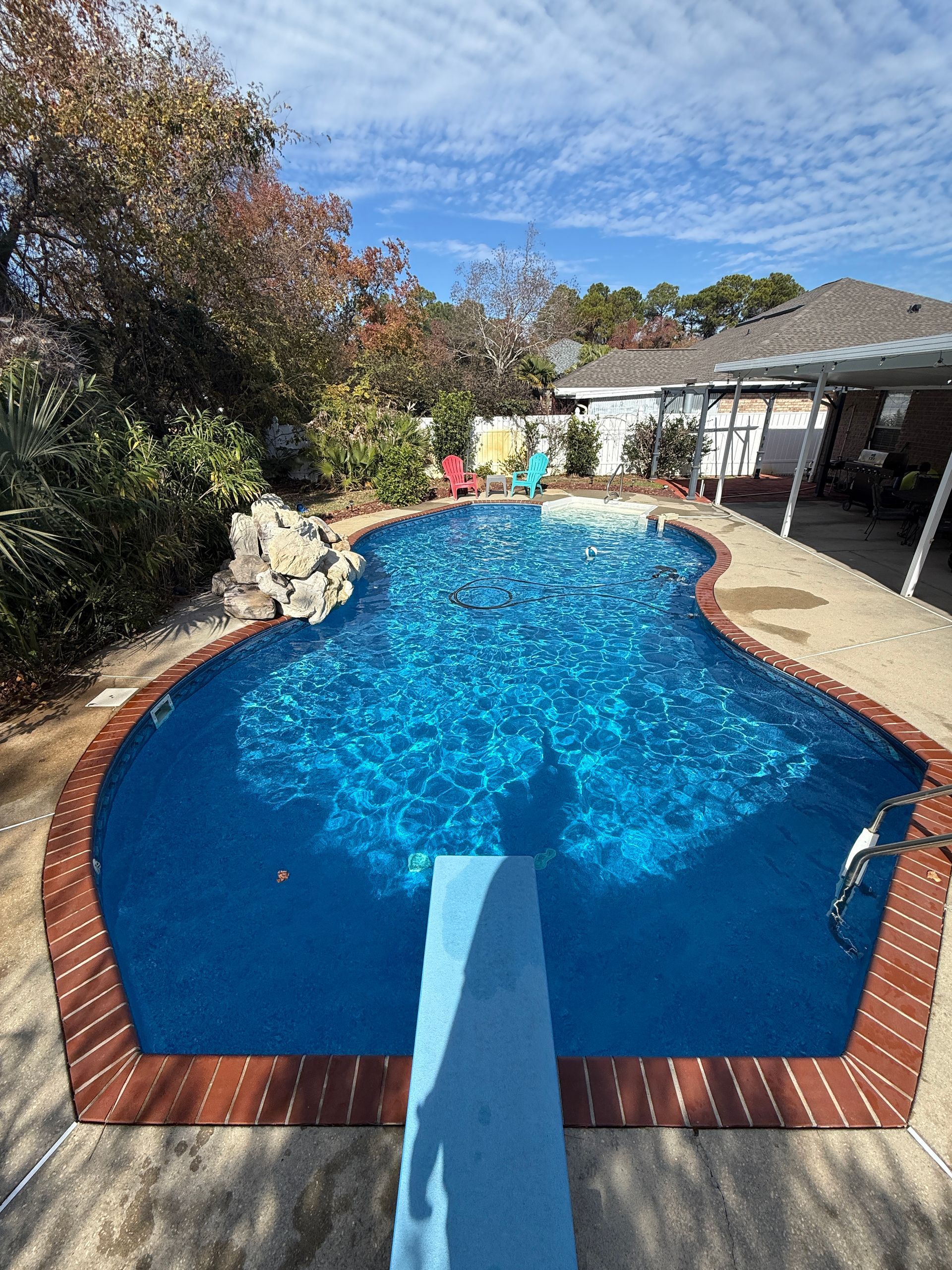 Blue swimming pool with a diving board, surrounded by a brown deck. Bright sunny day.