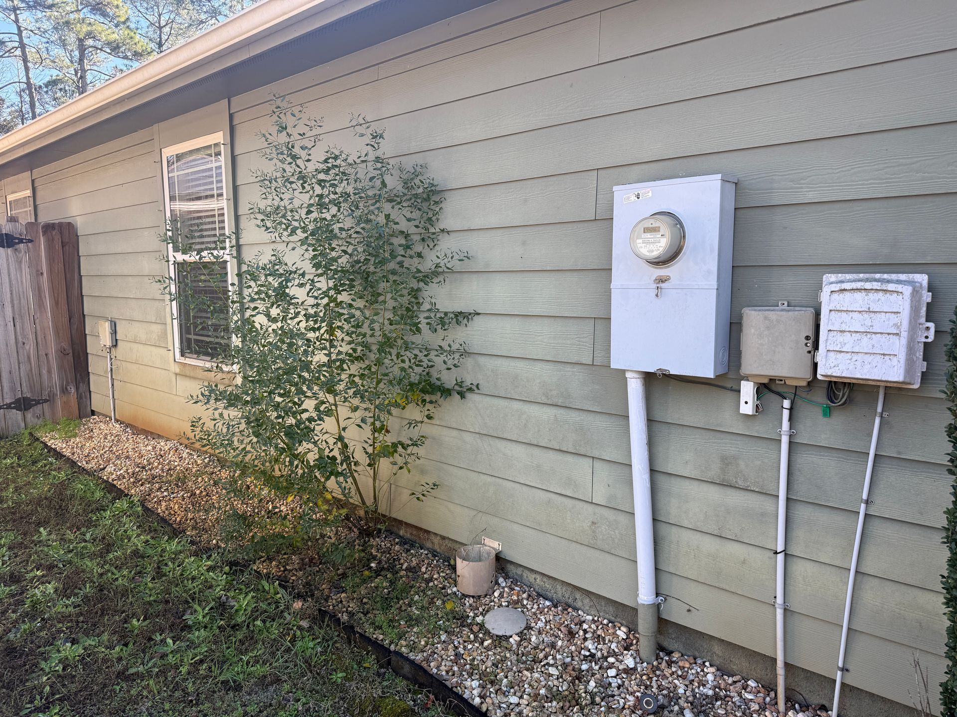 Exterior wall with electrical meter, boxes, window, and small tree. Gravel bed along the wall's base.