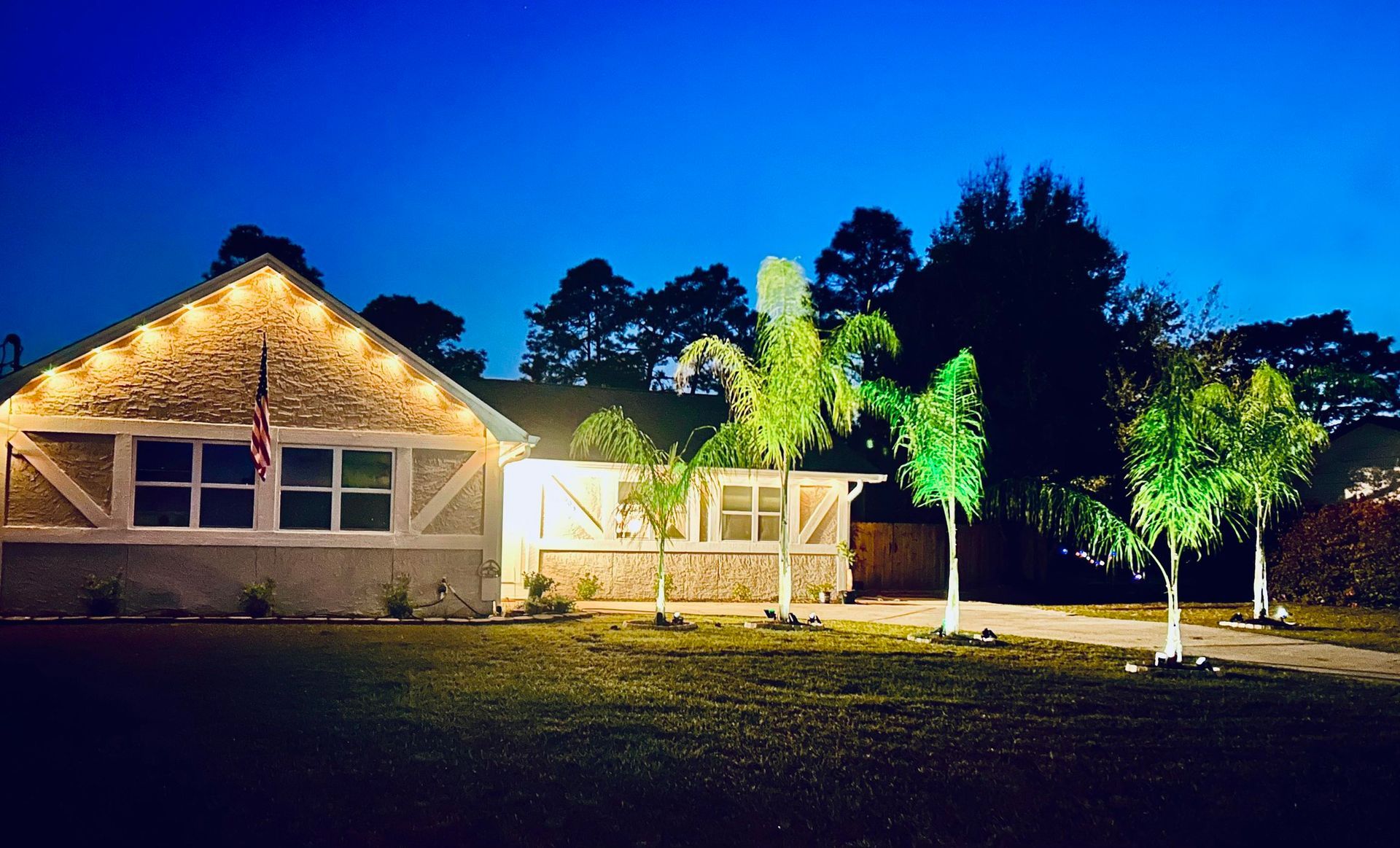 House at dusk, decorated with string lights, lit palm trees in front.