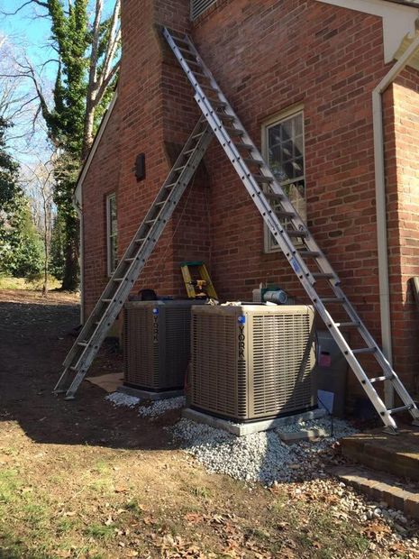 Two ladders leaned against a brick house beside HVAC units on a gravel pad outdoors
