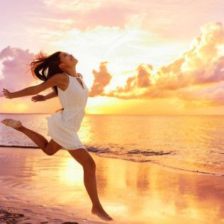 Woman in white dress leaping joyfully on a beach at sunset.