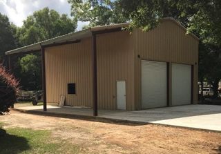 Tan metal storage building with roll-up doors and a small side door on a concrete pad, surrounded by trees.