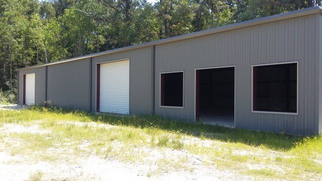 Gray metal warehouse with a white roll-up door and open bays, set beside trees and grass.