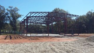 Steel building frame under construction on a cleared dirt lot with trees in the background