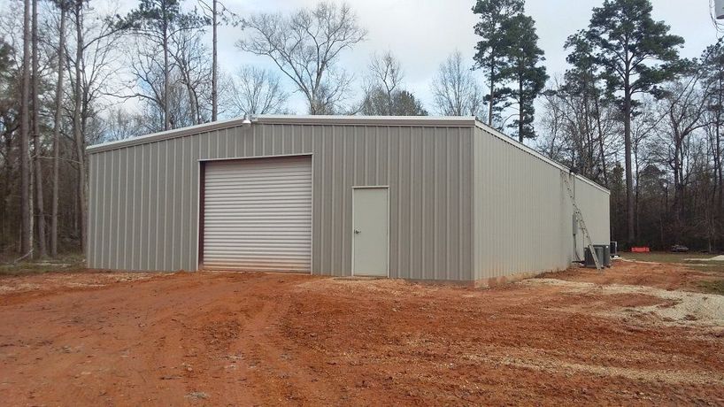 Large gray metal storage building with a roll-up door on a dirt lot surrounded by trees
