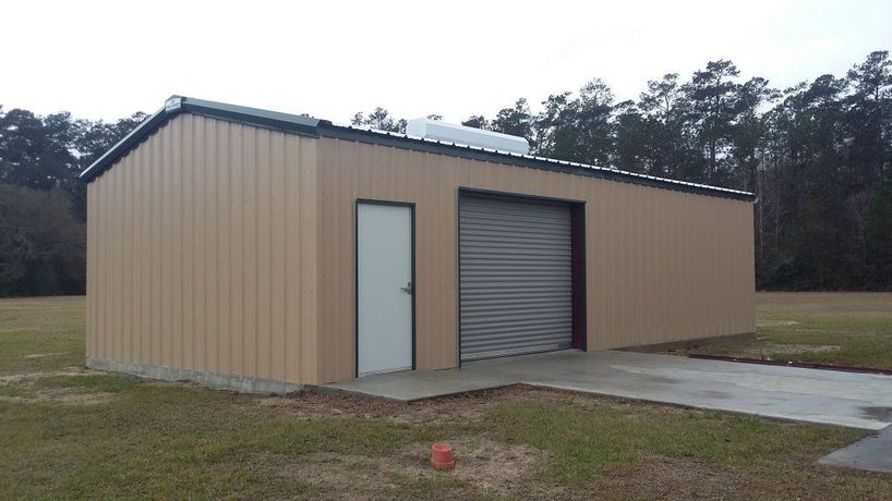 Tan metal storage building with white door and roll-up garage door beside a concrete driveway.