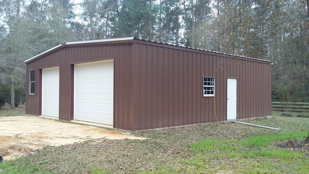 Brown metal storage building with white roll-up garage doors in a wooded clearing