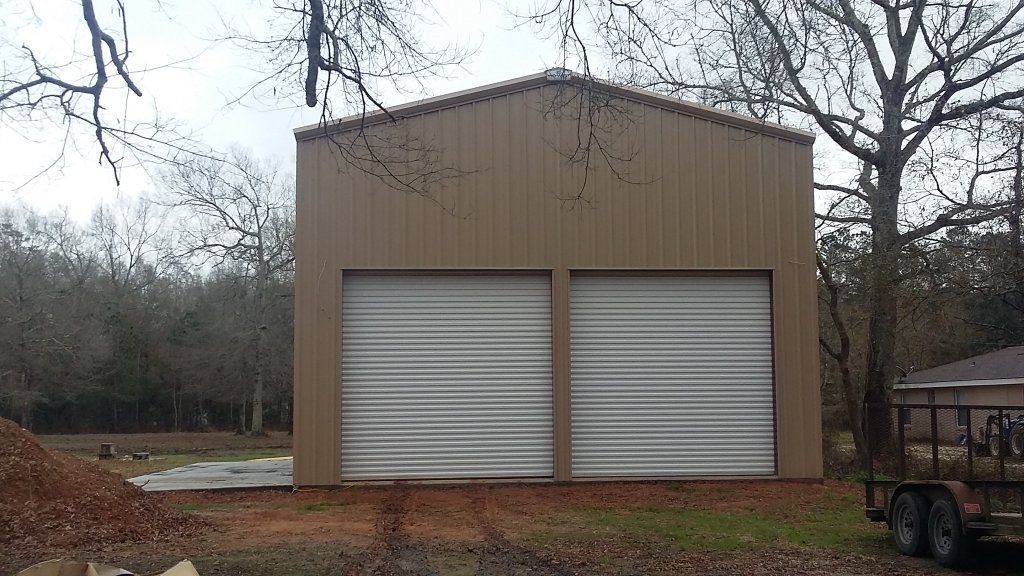 Tan metal storage building with two white roll-up garage doors in a wooded area.