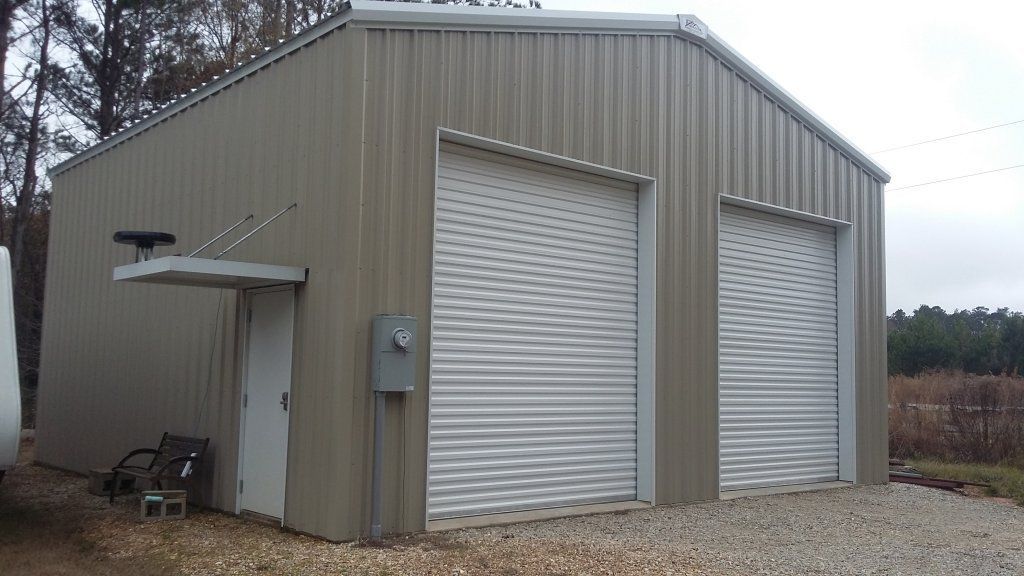 Gray metal garage or workshop with two roll-up doors and a small side awning on a gravel lot