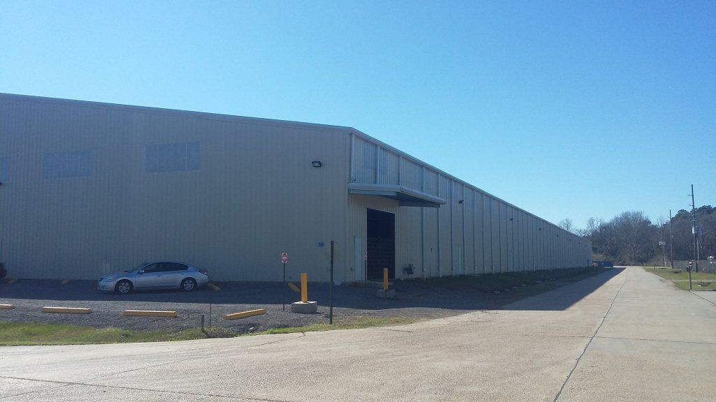 Large beige warehouse building with a parked car and a wide concrete driveway under a clear blue sky
