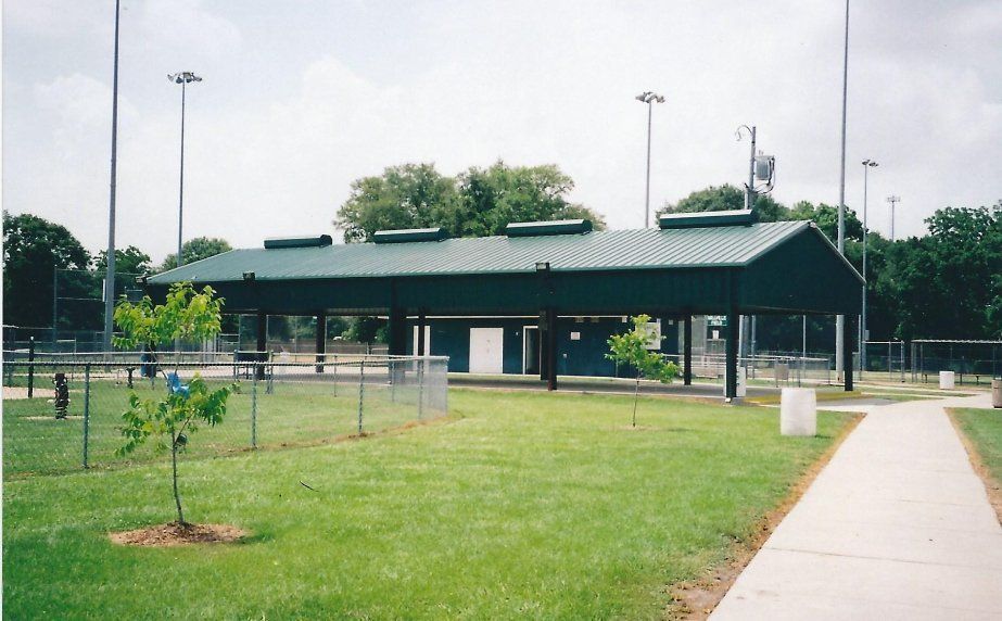 Covered picnic shelter in a park beside a sidewalk, with grass, chain-link fence, and trees.