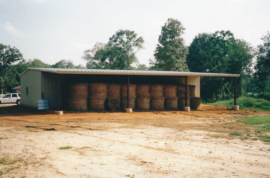 Rural storage shed with stacked hay bales under a tin roof, set on a dirt lot with trees behind.
