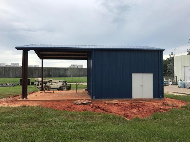 Blue metal shed with open canopy on a red dirt lot beside a grassy field