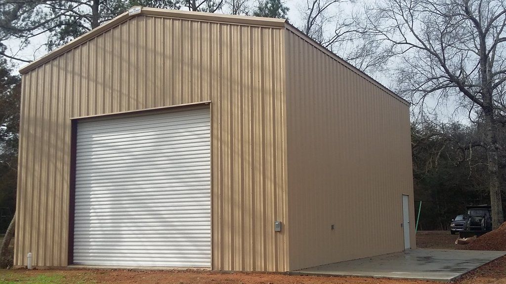 Tan metal storage shed with a white roll-up garage door beside leafless trees.