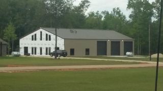 White building with black garage doors beside a dirt road, surrounded by trees and grass.
