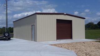 Tan metal storage building with a red roll-up door on a concrete pad under a blue sky