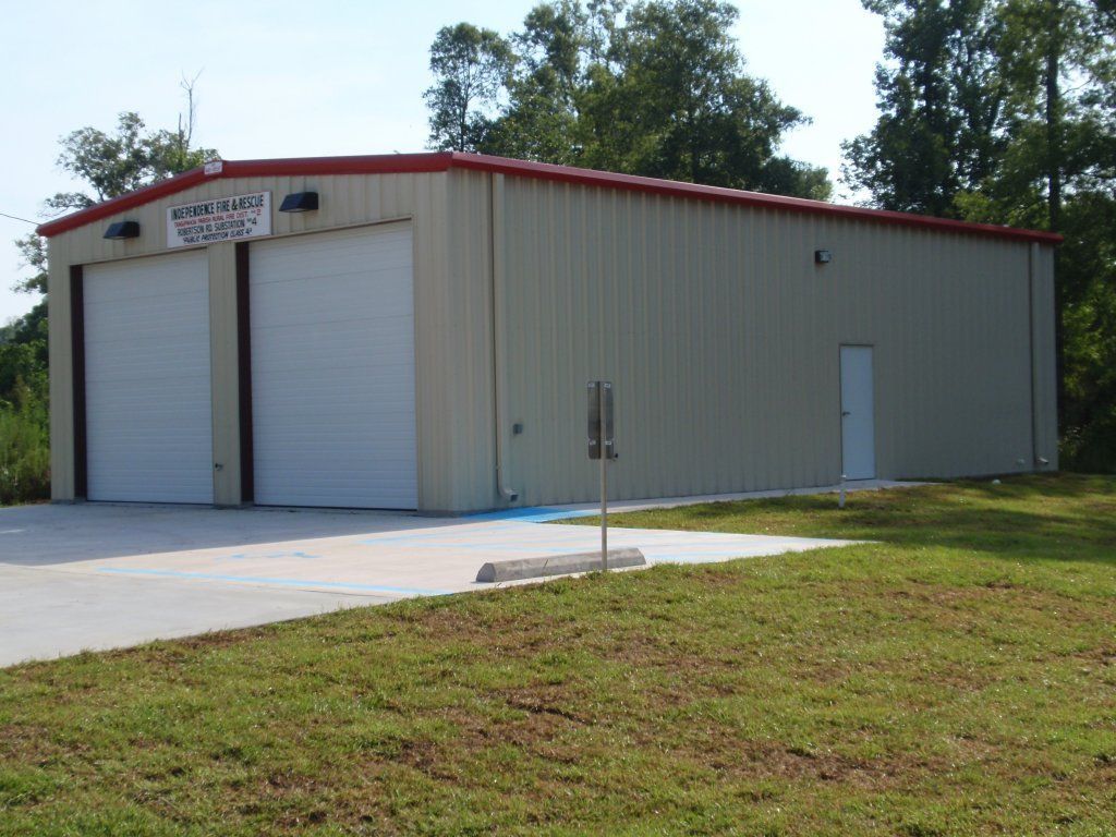 Small beige metal warehouse with red trim, roll-up doors, and a concrete driveway beside a grassy lot.