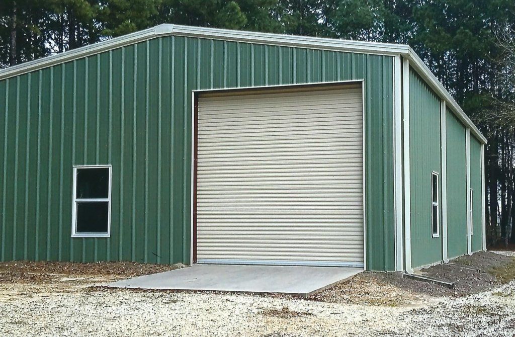 Green metal storage building with a large white roll-up door and two windows on a gravel lot
