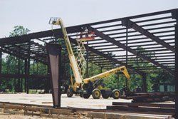 Crane lifting steel beams at a construction site with a large metal frame structure.