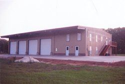 Brown warehouse with three garage doors and a side entry, viewed across a grassy lot at dusk