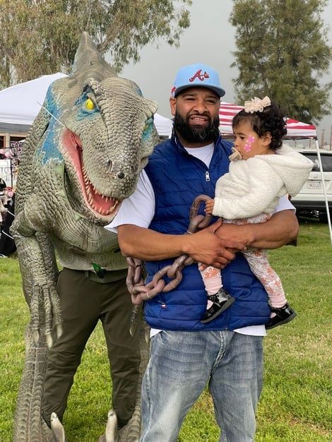 Man posing with a child beside a life-size dinosaur at an outdoor event