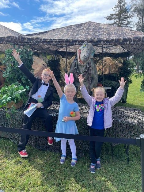 Three children posing with dinosaur statues outdoors, one holding a small orange egg and others wearing bunny ears.
