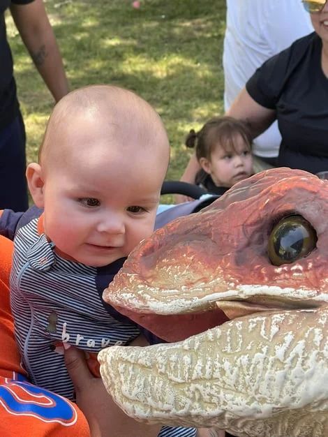 Baby reaching toward a dinosaur model at an outdoor event, with adults nearby.
