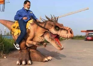 Man riding a realistic dinosaur costume outdoors near a red car.