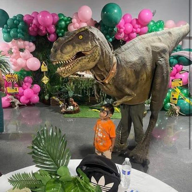 Child in orange shirt posing beside a large dinosaur statue, with pink and green balloon decorations behind.