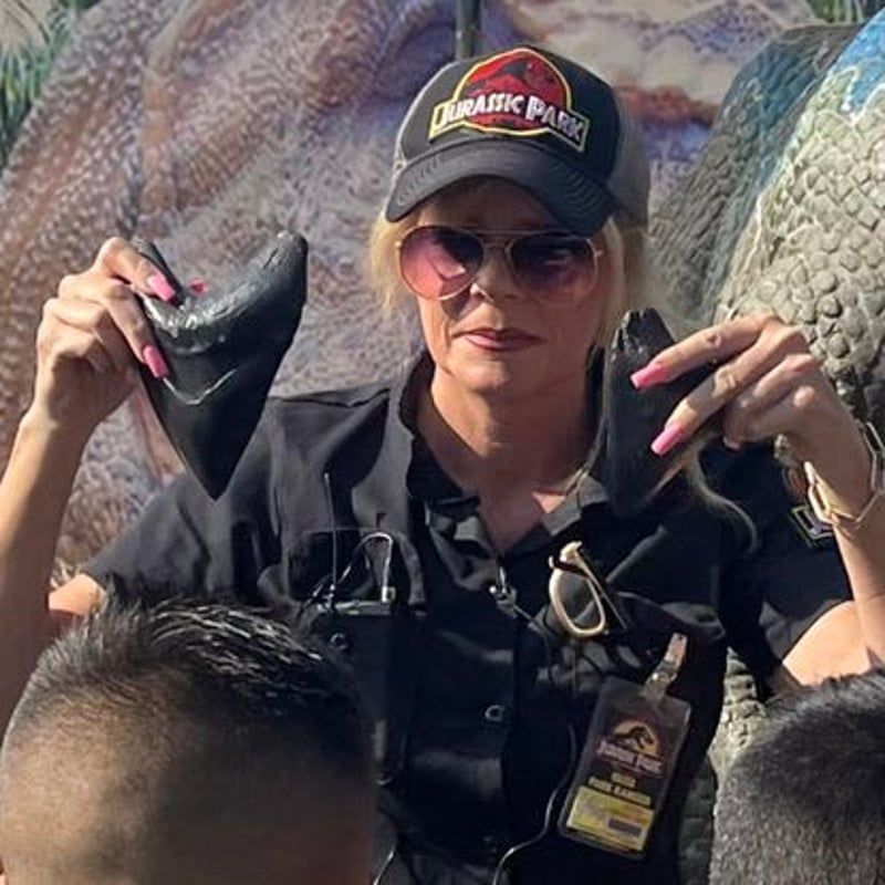 Person in black hat and sunglasses holding up two black shoes at an outdoor event, with dinosaur backdrop.