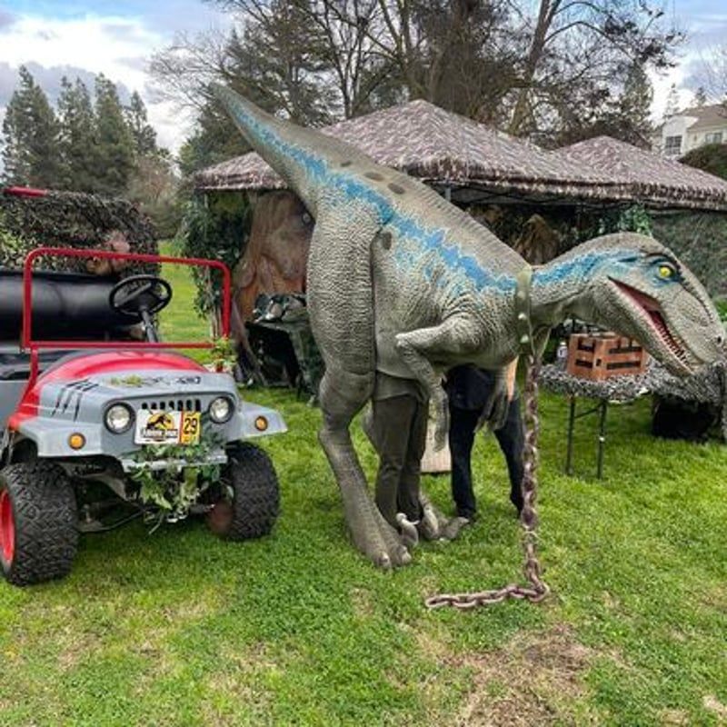 Gray dinosaur statue beside a red utility cart on a grassy lawn in front of a house