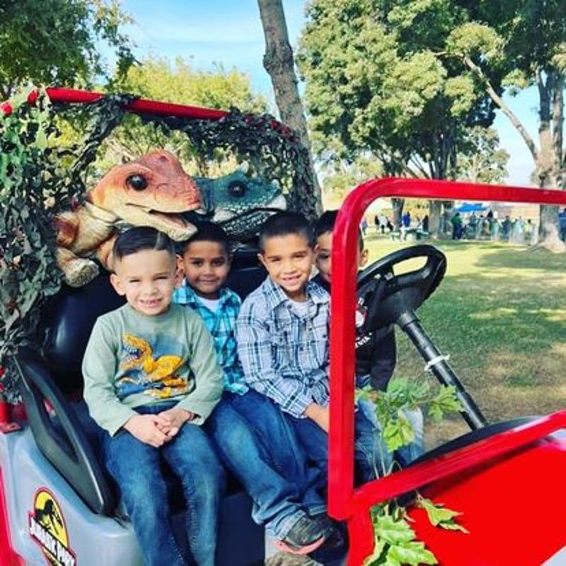 Three children smiling in a red vehicle at a dinosaur-themed park display
