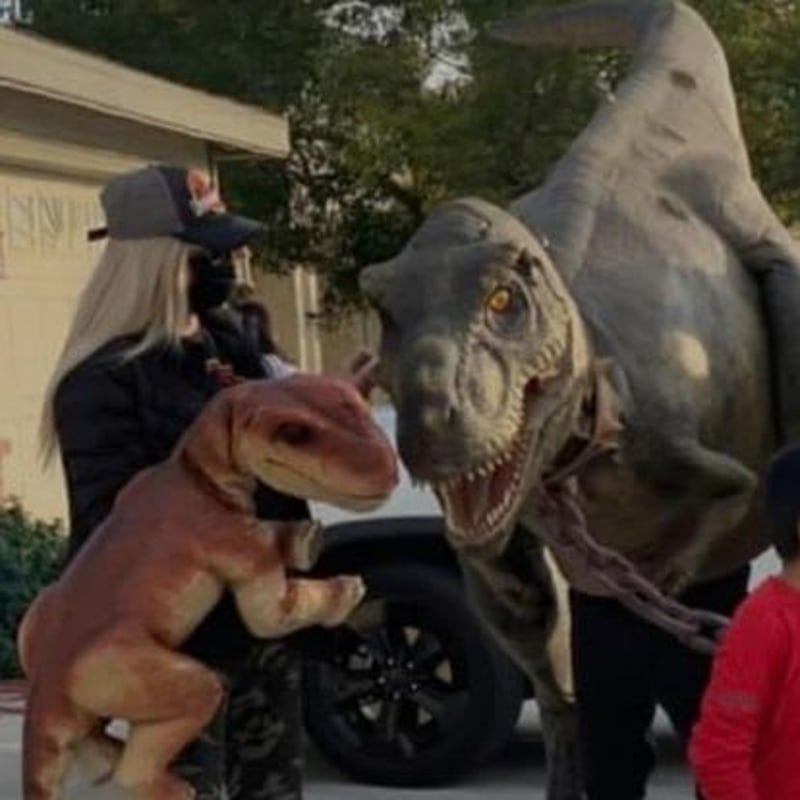 Two dinosaur costumes facing each other outdoors beside a parked car, with a person nearby.