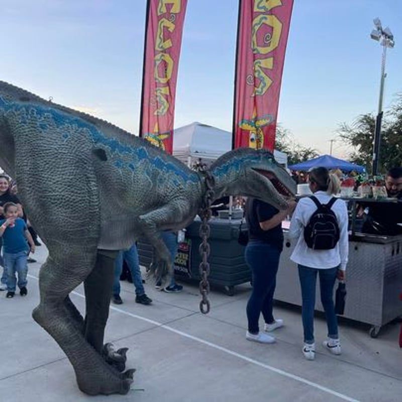 Large blue dinosaur statue beside people at an outdoor fair with colorful banners
