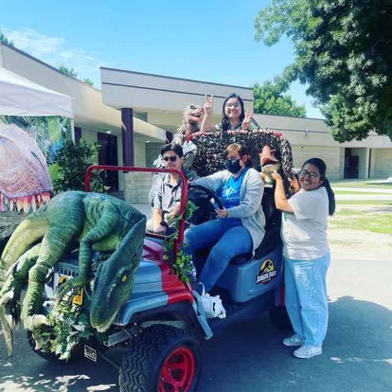 Group posing on a decorated golf cart with a large green dinosaur in front, outdoors on a sunny day