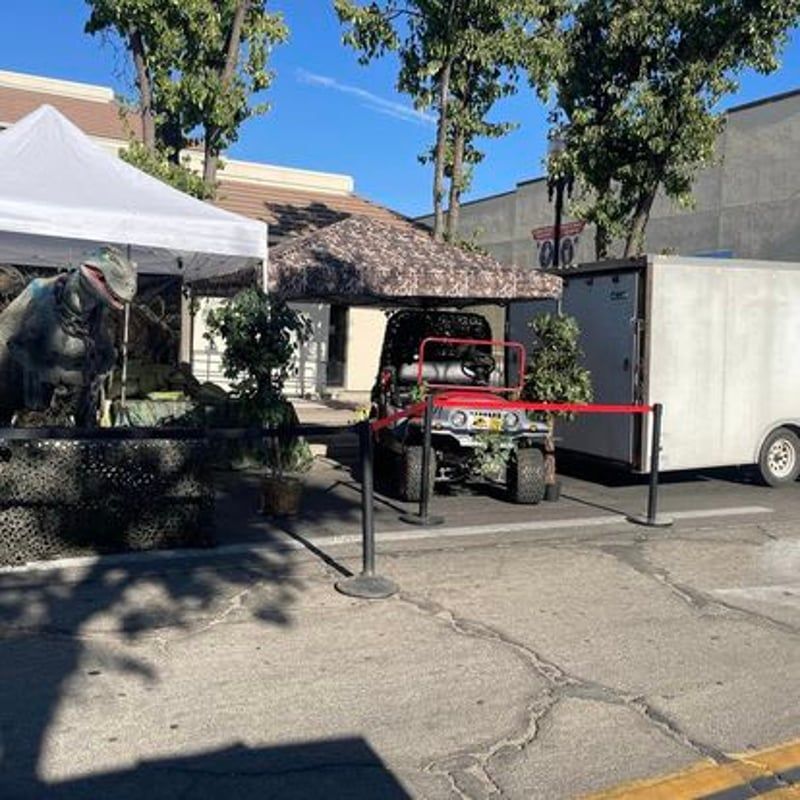 Outdoor event booth with a dinosaur display and a red utility vehicle behind a barrier rope