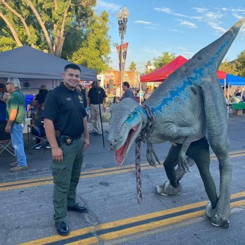 Man standing beside a blue-gray dinosaur costume at an outdoor street festival