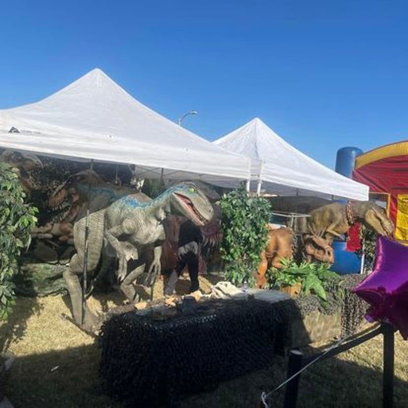Dinosaur sculptures under white tents at an outdoor fair, with a black table in front and blue sky overhead