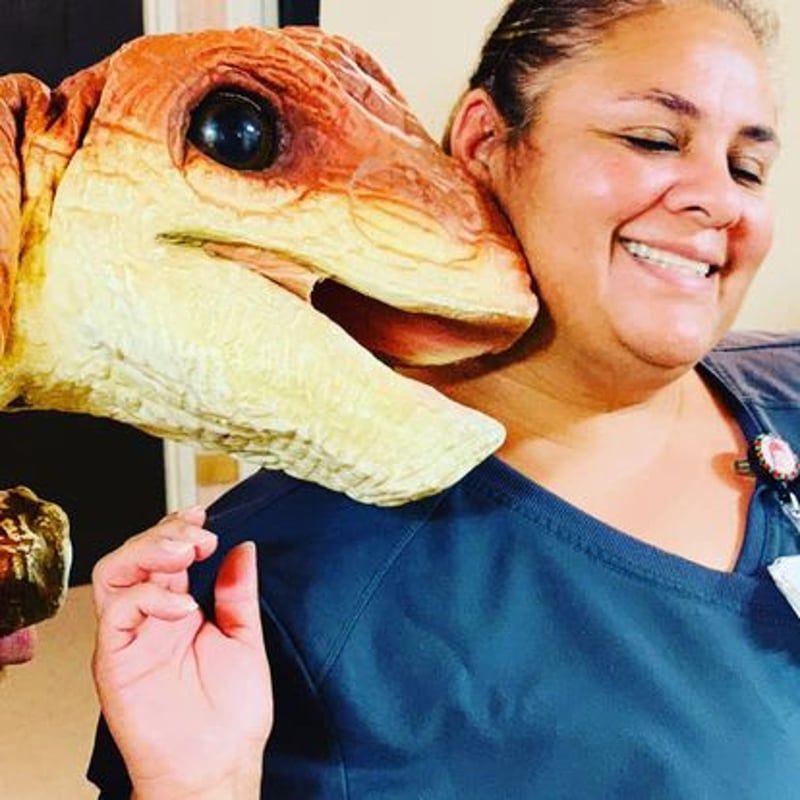 Woman smiling beside a large dinosaur puppet head with an orange face and black eye