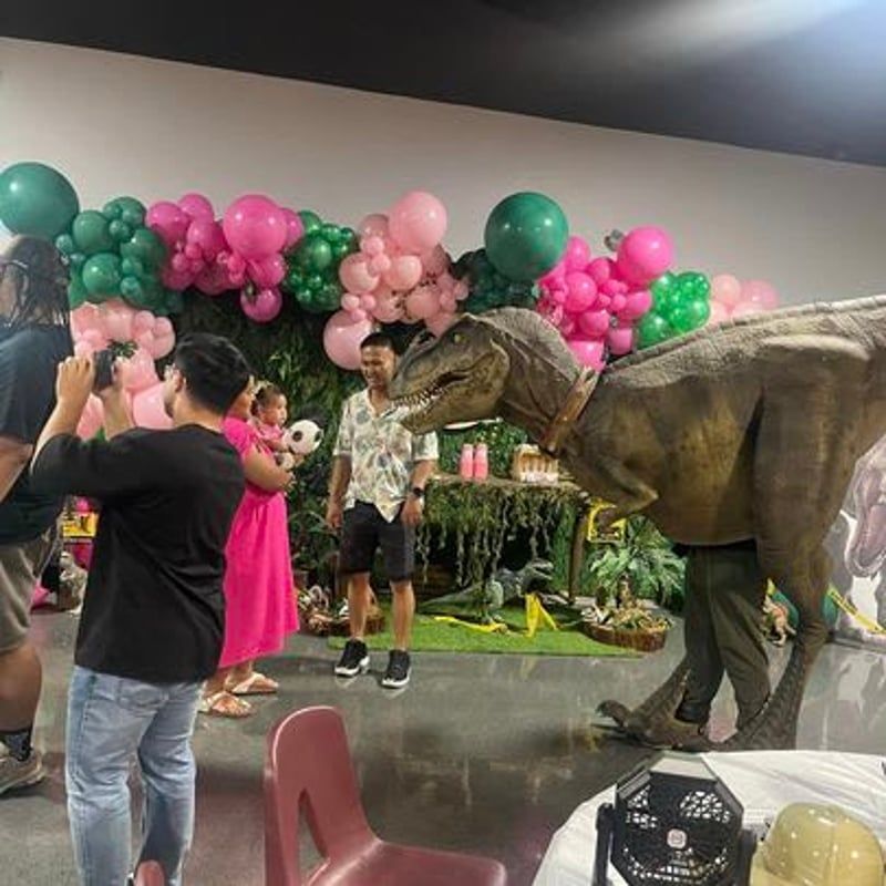 T-Rex model at a party with pink and green balloon garlands and people posing nearby