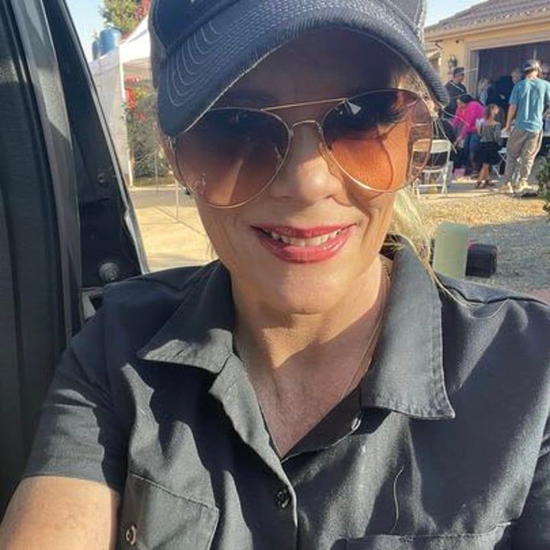 Smiling person in a cap and sunglasses taking a selfie outdoors in a dark shirt.