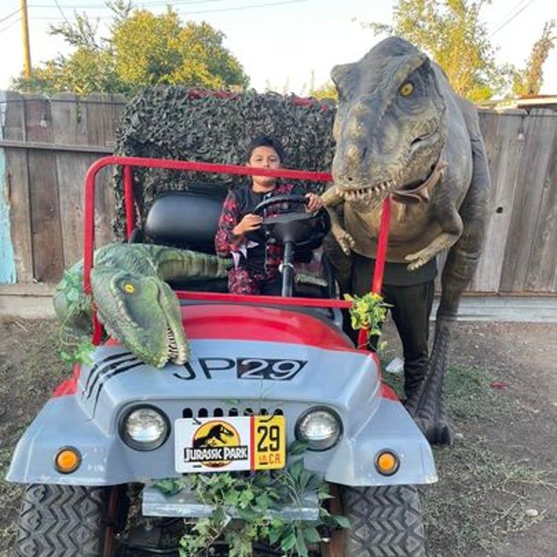 Child in a toy car with two dinosaur figures, outdoors by a wooden fence.