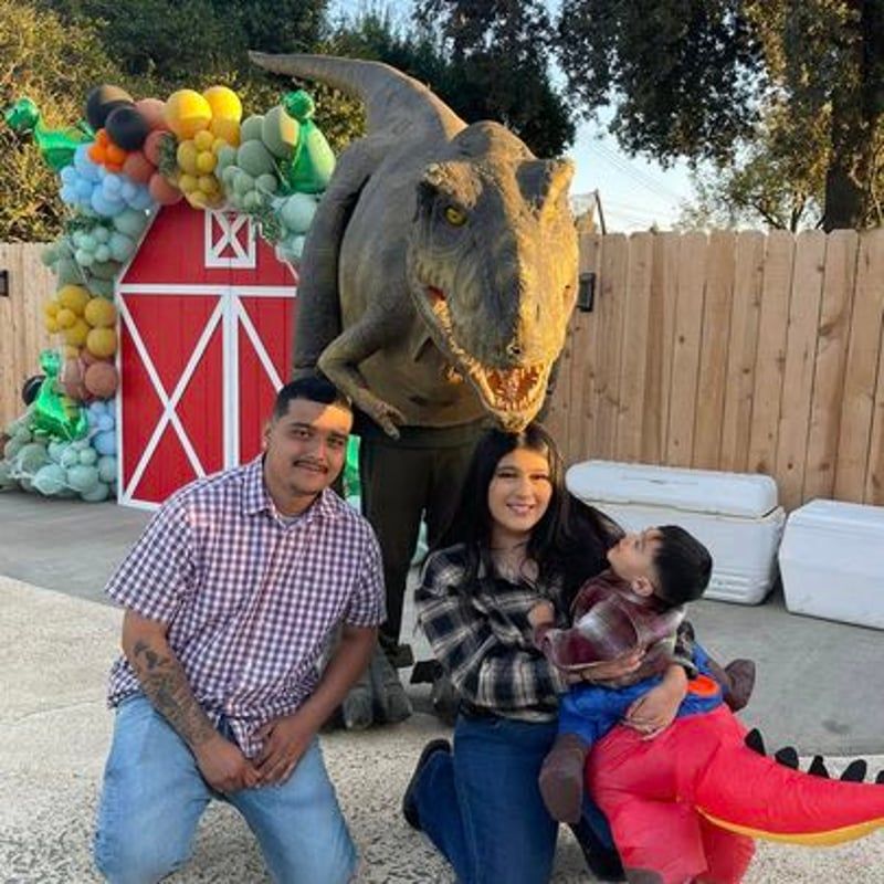 Family posing beside a large dinosaur statue and colorful balloon barn backdrop outdoors