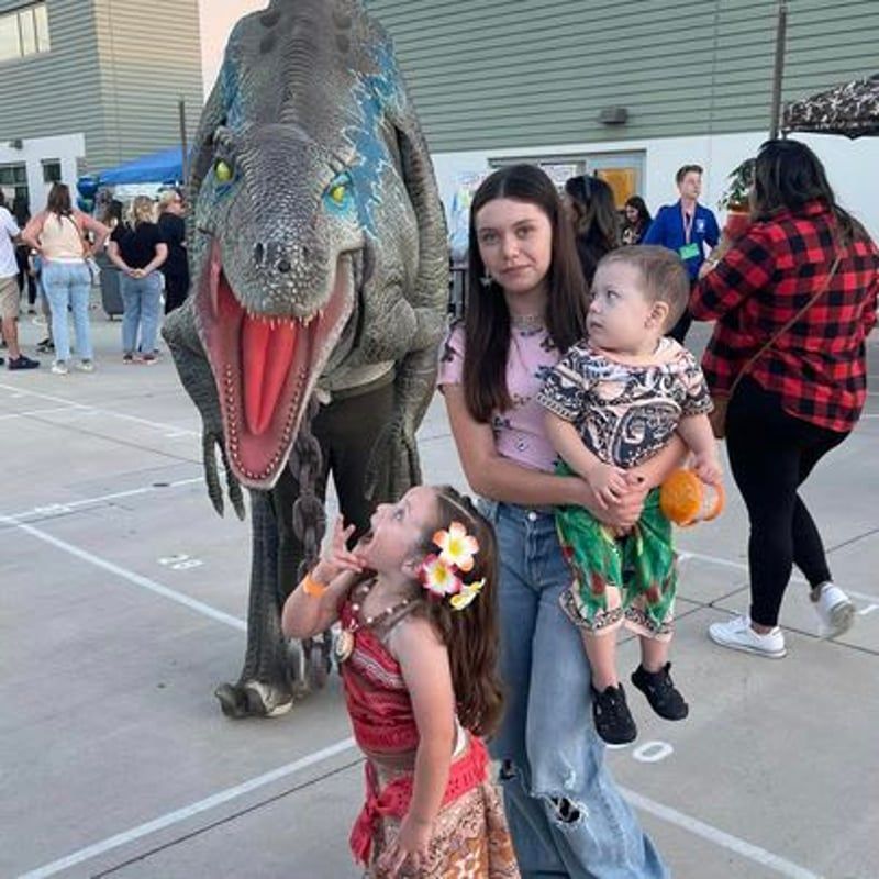 Person in dinosaur costume at an outdoor event, with children nearby and a woman holding a toddler