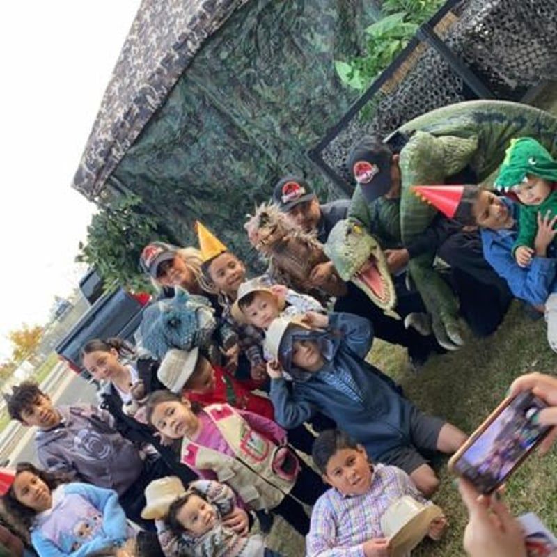 Costumed group posing outdoors, including dinosaurs, a shark, and a dragon, with a phone capturing the moment